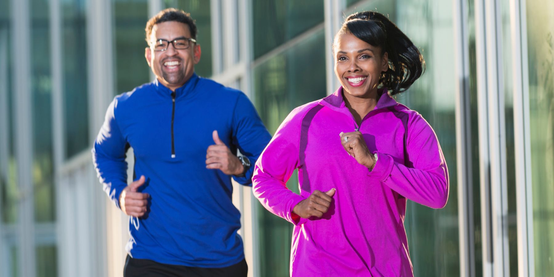 A happy, mature, African American couple, in their 40s, jogging or running on a city sidewalk, next to an office building. The woman is in focus, running in front, with the man a few steps behind. They are wearing colorful sweatshirts, purple and royal blue, and dark gray pants. They are both smiling.
